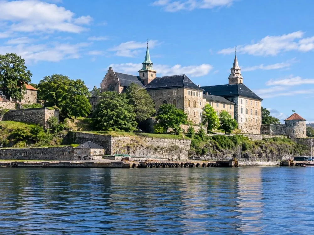 Akershus Fortress seen from the fjord: pale stone walls and steep roofs standing on a green headland directly above the harbour water, with the quayside below the ramparts.