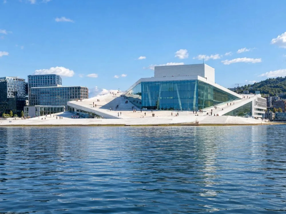 The Oslo Opera House across Bjørvika: a low white building of pale stone and glass sloping straight down into the water, with people small on its sloping roof.
