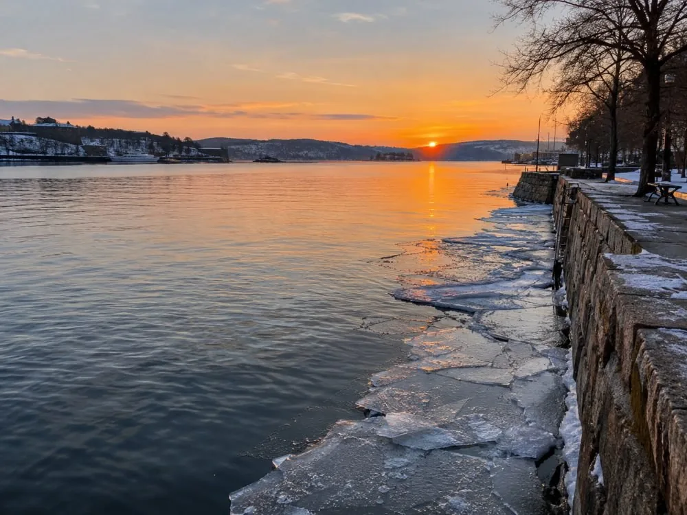 The inner Oslofjord on a winter afternoon: flat grey water, thin ice at the edge of a quay, and the low orange light of a sun that will set in the middle of the afternoon.